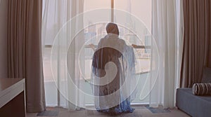 Back view of woman opening curtains and looking out of window to cityscape at hotel in the morning. Modern buildings at