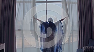 Back view of woman opening curtains and looking out of window to cityscape at hotel in the morning. Modern buildings at