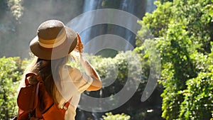 Back view of woman with photo camera shooting tropical waterfall. Inspiring millennial travel blogger in journey. Happy