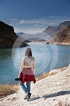 Back view a woman photographer in casual clothes with a professional camera stands on the shore of a lake in the