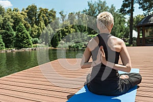 back view of woman practicing yoga in lotus pose and hands in namaste gesture on yoga mat near river