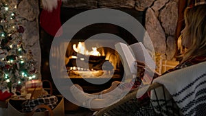 Back view woman reading book and relaxing at cozy burning fireplace on Christmas