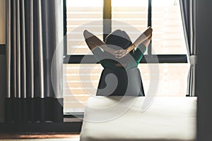 Back view of woman relaxing and sitting on chair with hands on her head and looking on window in bedroom at home