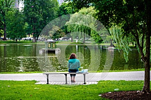 Back view of a woman sitting alone on a bench facing a lake in Boston Commons public park