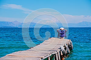 Back view of a woman sitting on a tiny wooden pier by the sea. Young beautiful girl sitting on jetty. Back view. Amazing