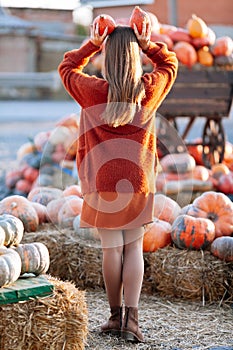 Back view of woman with small pumpkin in hand above head near wooden wagon with pumpkin on farmers market in brown sweater. Cozy