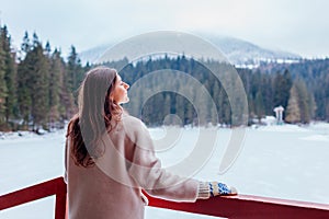 Back view of woman tourist admiring snowy mountain Synevyr lake view wearing white warm fur coat. Winter landscape photo