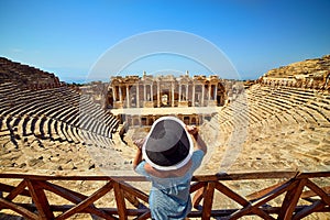 Back view of woman traveler in hat looking at amazing Amphitheater ruins in ancient Hierapolis, Pamukkale, Turkey. Grand panorama