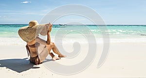 Back view of woman on tropical vacation relaxing on the sandy beach with turquoise water, wearing bikini and summer hat. Paradise