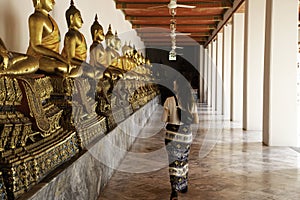 Back view of a woman walking side to aligned golden Buddha Statues at the Emerald temple in Bangkok