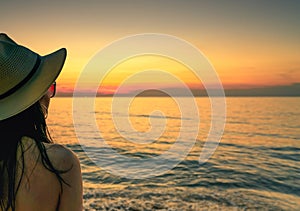 Back view of woman watching sunset at the beach. Woman wear sunglasses and straw hat relaxing at tropical paradise beach.