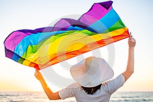 Back view of woman in white dress and hat holding gay pride flag at sunset in beach