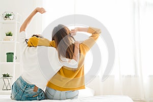 Back view of women lesbian happy couple waking up in morning, sitting on bed, stretching in cozy bedroom, looking through window.