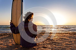 Back view young adult slim sporty female surfer girl with surfboard sitting on sand at ocean coast wave against warm