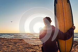 Back view young adult slim sporty female surfer girl with surfboard sitting on sand at ocean coast wave against warm