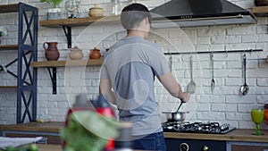 Back view of young Asian man mixing food in pan in slow motion. Guy cooking healthful breakfast in kitchen in the