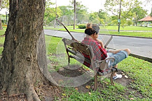 Back view of young asian man in red shirt reading a book on the bench against in beautiful outdoor park.