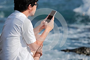 Back view of young Asian man using mobile smart phone at sea shore. Internet of things concept