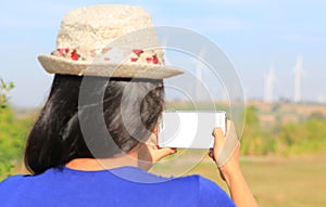 Back view of young Asian woman taking photo with smartphone of wind turbine field