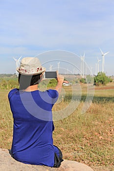 Back view of young Asian woman taking photo with smartphone of wind turbine field