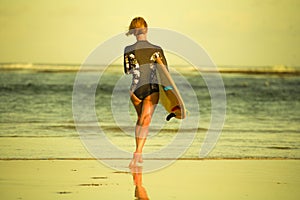 Back view of young attractive and sporty surfer girl in cool swimsuit at the beach carrying surf board into the sea running toward
