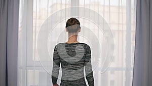Back view of young attractive woman opening curtains on a big window and letting the light in the room. Looking out the