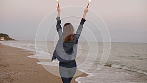 Back view of young attractive woman running by the sea during sunset and holding burning sparkling candles in both hands