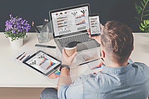 Back view.Young bearded businessman in denim shirt sits in office at table and uses smartphone with charts,diagrams