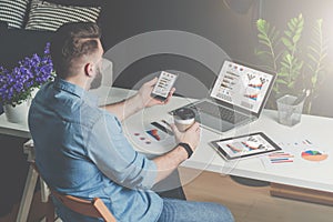 Back view.Young bearded businessman in shirt sits in office at table and uses smartphone with charts,diagrams and graphs