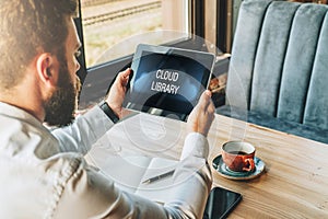 Back view. Young bearded businessman is sitting at table, using tablet computer with inscription on screen- cloud library