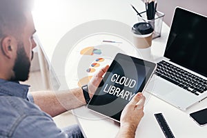 Back view. Young bearded businessman is sitting at table, using tablet computer with inscription on screen- cloud library