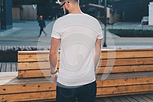 Back view. Young bearded hipster man dressed in white t-shirt and sunglasses is stands on city street. Mock up.