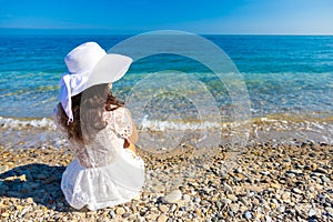 The back view of young beautiful woman in white dress sitting on the beach and looking at the sea