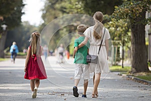 Back view of young blond long-haired woman walking with two children, small daughter and son along sunny park alley on blurred