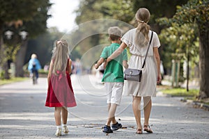 Back view of young blond long-haired woman walking with two children, small daughter and son along sunny park alley on blurred