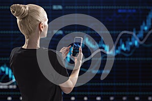 Back view of young blonde woman using mobile phone with glowing candlestick forex chart and index on dark grid background. Trade,