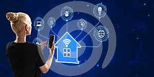 Back view of young blonde woman using phone with creative smart house hologram on blue background. Artificial intelligence and
