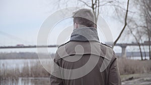 Back view of young brunette Caucasian man walking along the riverbank. Camera following thoughtful handsome guy