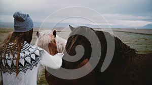 Back view of young brunette woman stocking two beautiful Icelandic horses grazing on a field in overcast day.