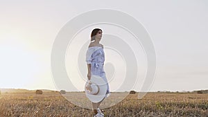 Back view. A young brunette woman walking a trail in the field at sunset. Attractive girl in a long blue dress with hat