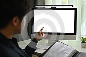 Back view of young businessman holding open office binder and pointing fingers at white blank screen computer monitor