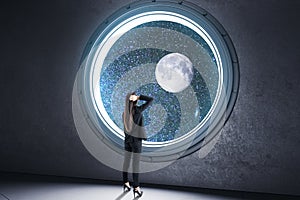 Back view of young businesswoman looking out of round illuminator with starry sky cosmos and planet view in concrete interior.