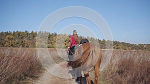 Back view of a young Caucasian female equestrian riding the black horse on the dirt road and holding halter of the brown