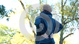 Back view Young Caucasian man massaging his back in green park between trees. Rear view sportsman stretching his body.