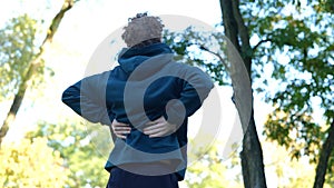 Back view Young Caucasian man massaging his back in green park between trees. Rear view sportsman stretching his body