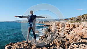 Back view of young caucasian man standing on edge of cliff with outstretched arms to sides. Attractive boy in blue shirt standing