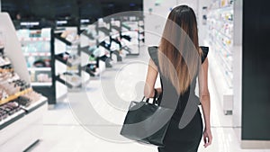 Back view of young Caucasian woman in black dress walking in cosmetics store.