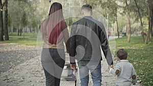 Back view of young couple walking with little son and baby stroller in spring or autumn park. Joyful Caucasian father