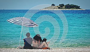 Back view of a young family sitting under an umbrella on a sunny beach in Croatia