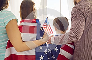 Back view of a young family standing at window wrapped in us flag celebrating Independence Day.